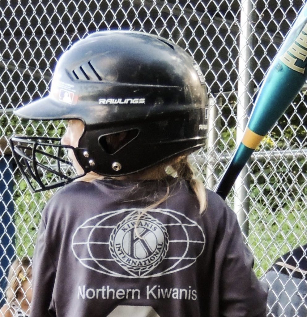 Girl with baseball bat and wearing helmet and a shirt with Kiwanis logo and Northern Kiwanis printed on shirt.