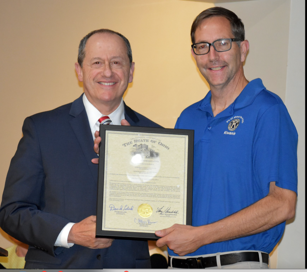 Kiwanis member in a blue shirt receiving resolution of congratulations from another man. dressed in a suit.